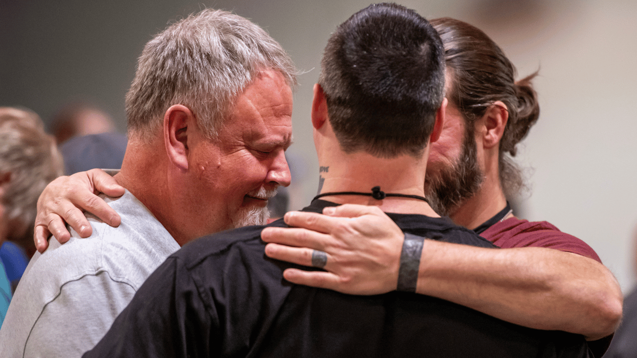 Three men hugging tightly, emotional, smiling, in an indoor setting with other people in the background.